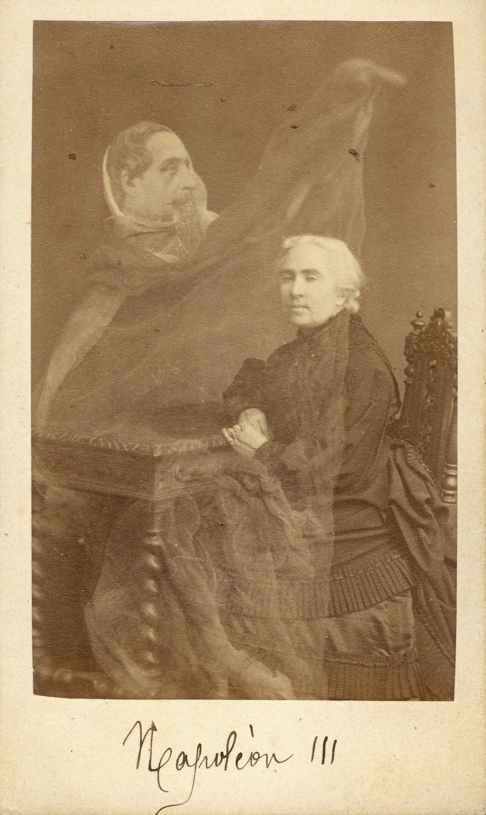 A carte-de-visite of a white-haired woman in black seated at a carved table, a translucent draped figure with a moustache and goatee behind her, one arm raised, and a name inked below.