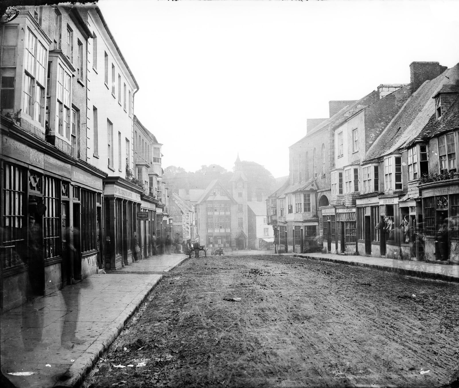 An old black-and-white view down a muddy, unpaved town street lined with shops and bay windows, with pale shapes on the left pavement and a horse and cart far down the road.