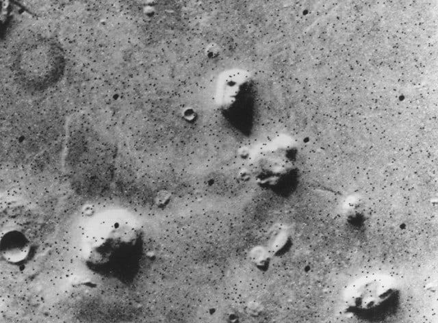 A grainy black-and-white overhead view of a pale, pitted plain peppered with black dots, with scattered mesas and craters and one rounded hill near the center.