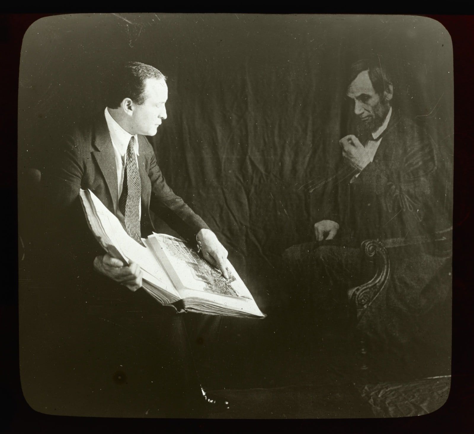 A lantern slide with rounded corners: a man in a suit holds a large open book at left, and a bearded man in a frock coat sits in a carved armchair at right.