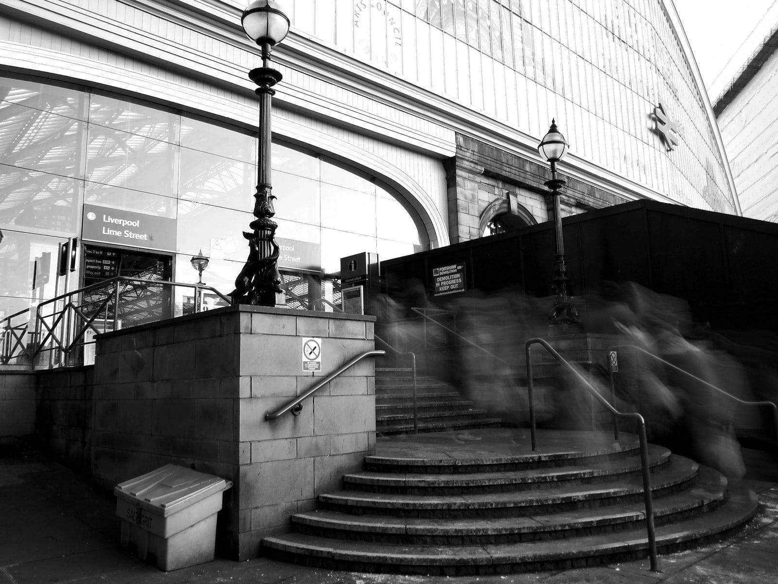 A black-and-white view of curved stone steps up to a railway station entrance, with ornate lamp posts, a Liverpool Lime Street sign and blurred grey shapes on the steps.