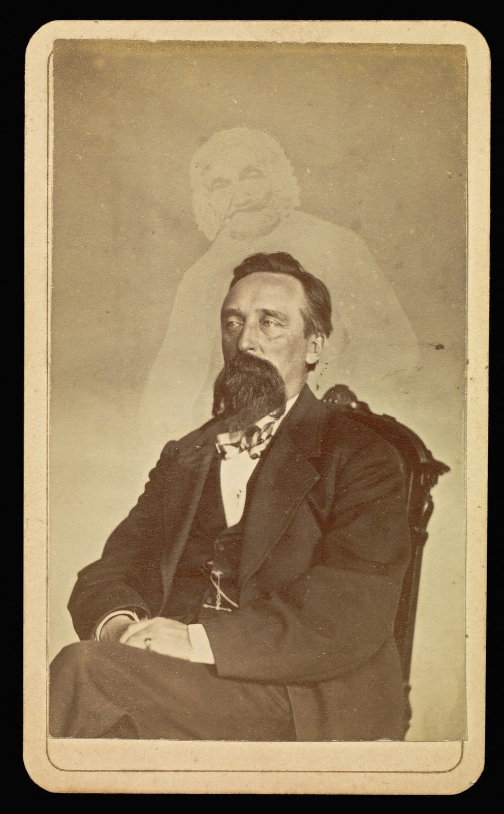 A carte-de-visite of a seated man with a large dark beard, a bow tie and a dark suit, looking left, with the pale head and shoulders of an old woman in a white cap above him.
