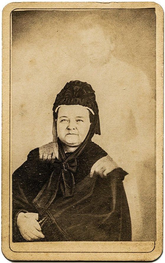A carte-de-visite portrait of an older woman in a black bonnet and cloak, seated, with a pale tall figure standing behind her, its hands on her shoulders.