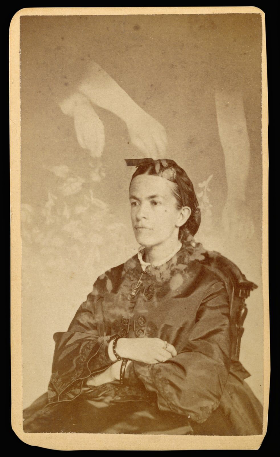 A carte-de-visite of a young woman in a dark, glossy dress, seated with her arms crossed, with pale arms, hands and faint sprays of flowers floating above her head.