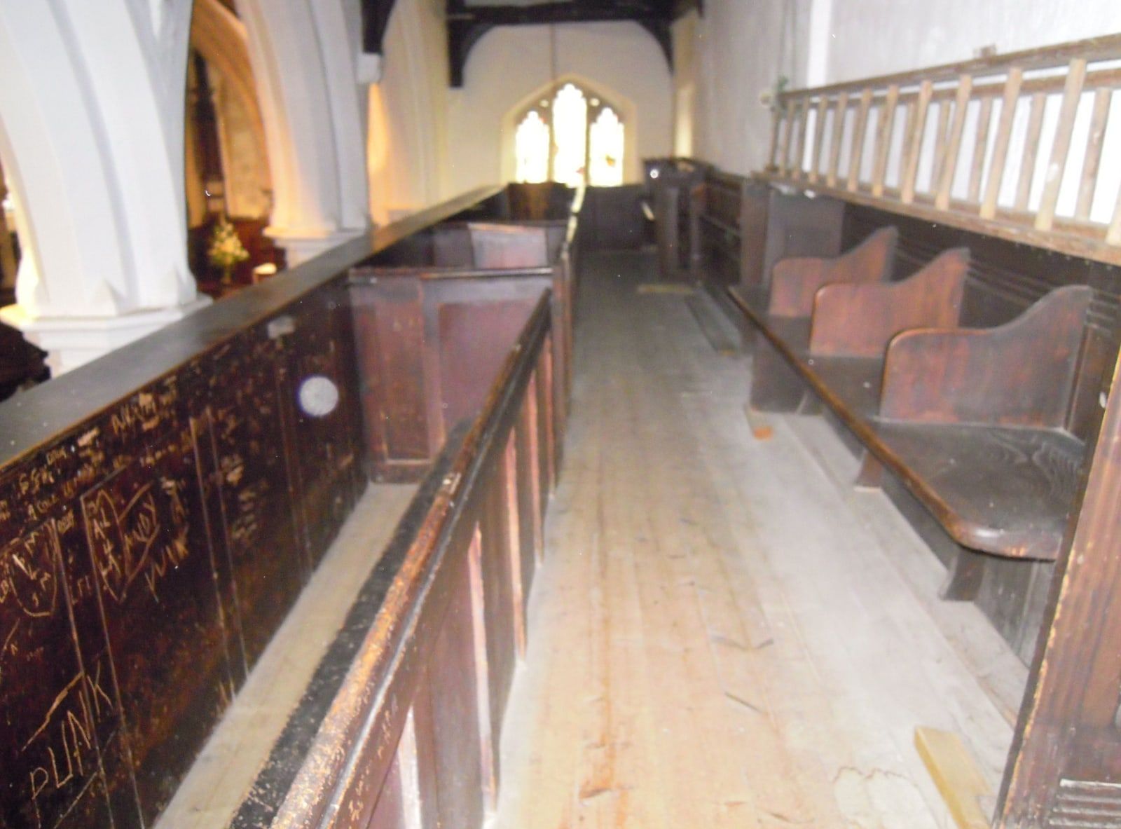 The wooden upper gallery of an old church: a plank floor, a dark panel covered in scratched graffiti on the left, old pews leading to an arched window, and a small pale circle.