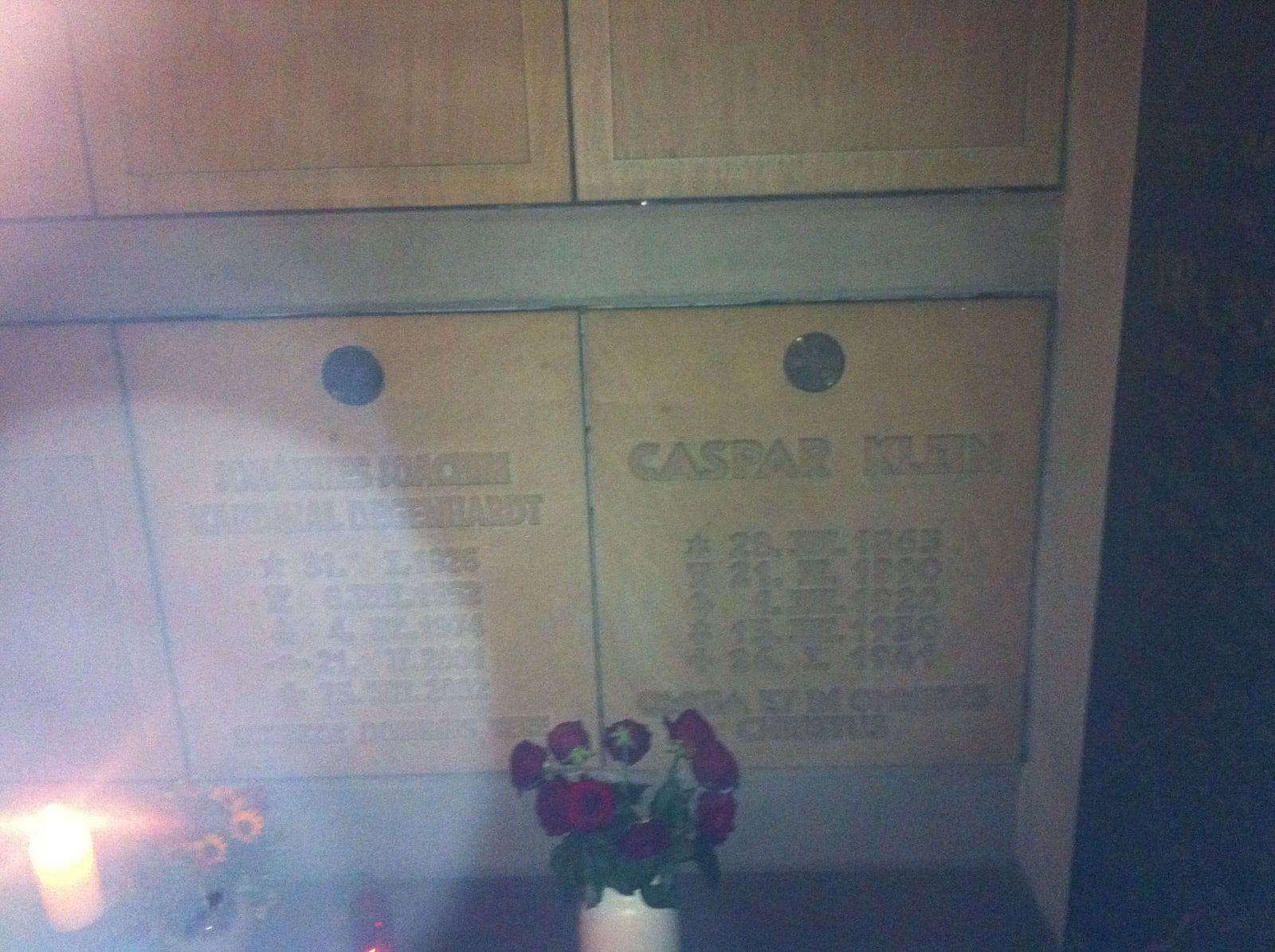 Stone burial niches in a dim crypt with engraved names and dates, red flowers in a vase and a lit candle at lower left, under a pale milky haze.
