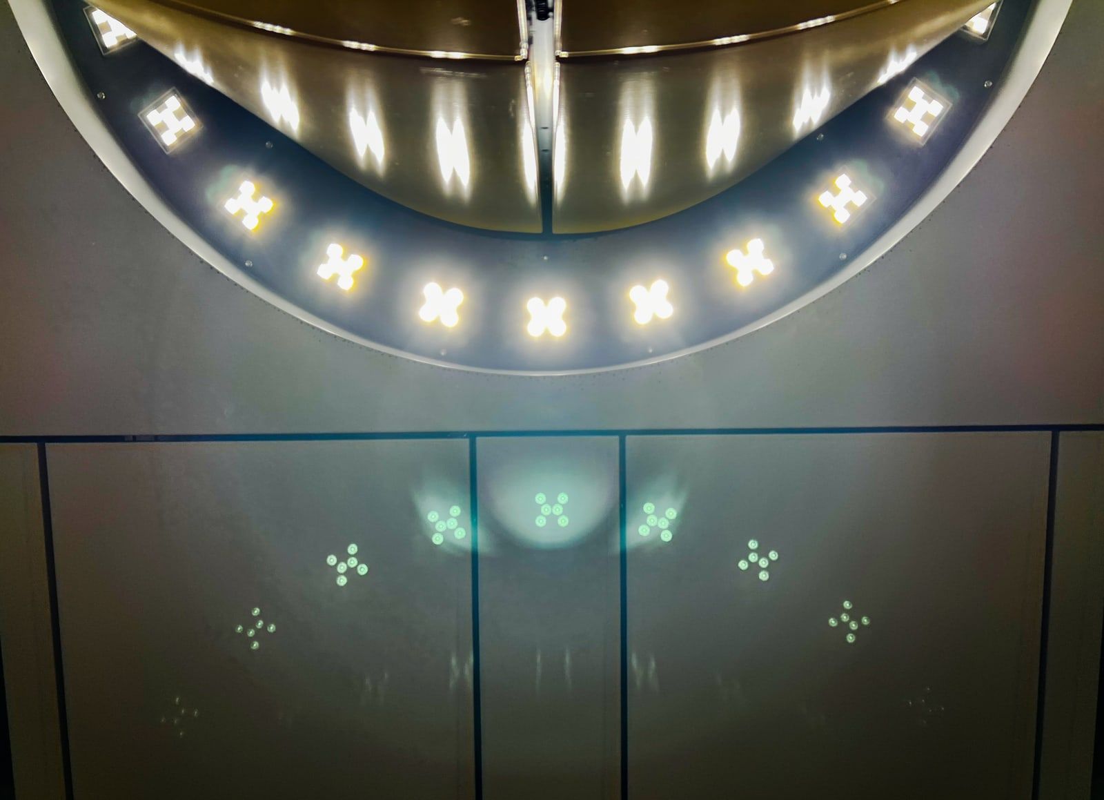 Looking up at a round ceiling light ringed with bright cross-shaped lamps above dark wall panels, with small green-white shapes and a pale glow floating on the panels below.
