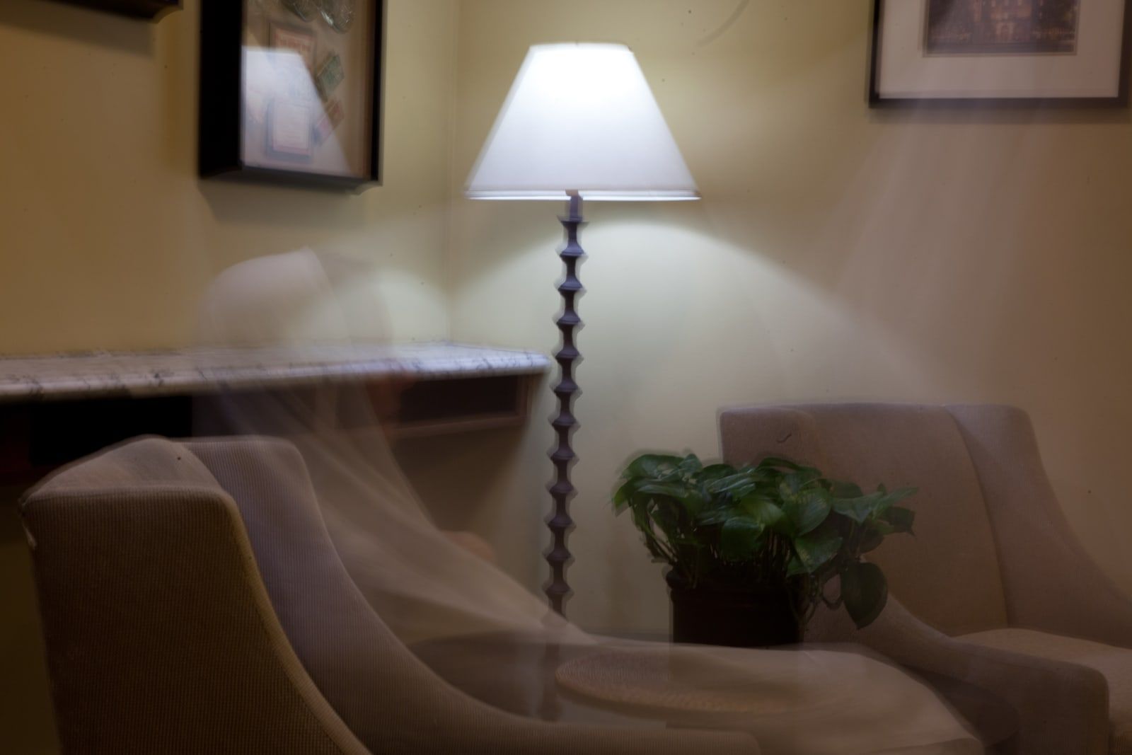 A quiet hotel corner with a floor lamp, a potted plant, two beige armchairs and a marble console, and a pale, see-through shape over the left armchair.