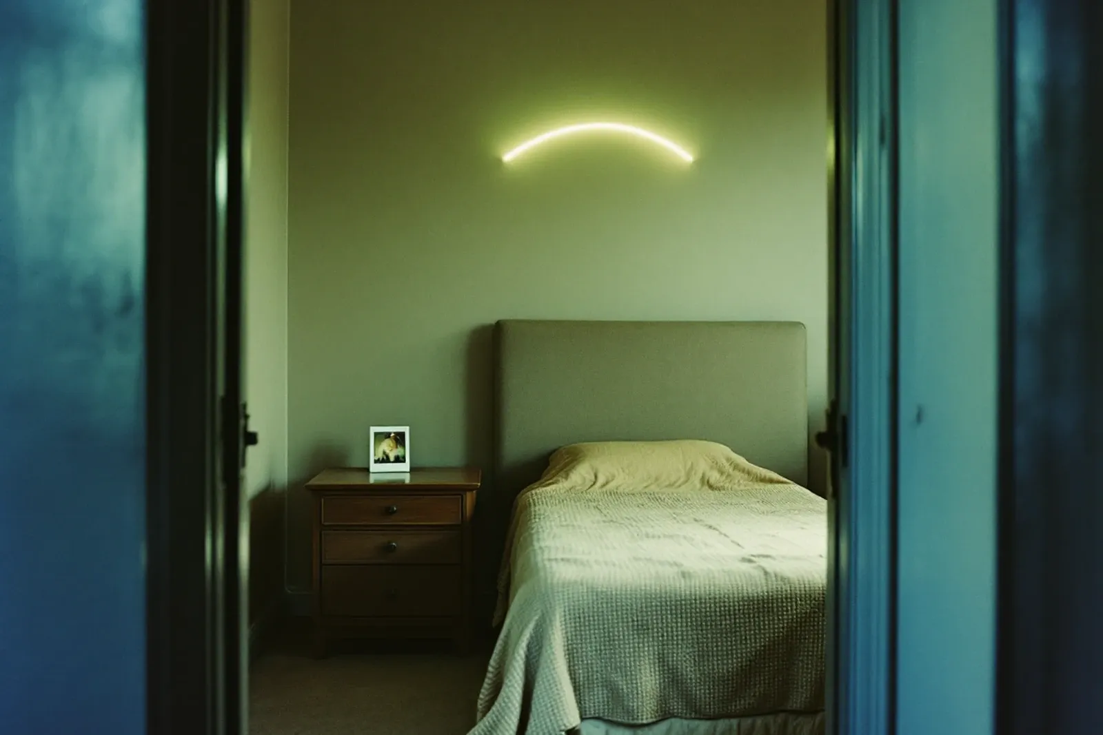 A Polaroid print face down on a bedside table in an empty 1970s bedroom, a pale arc of light on the wall.