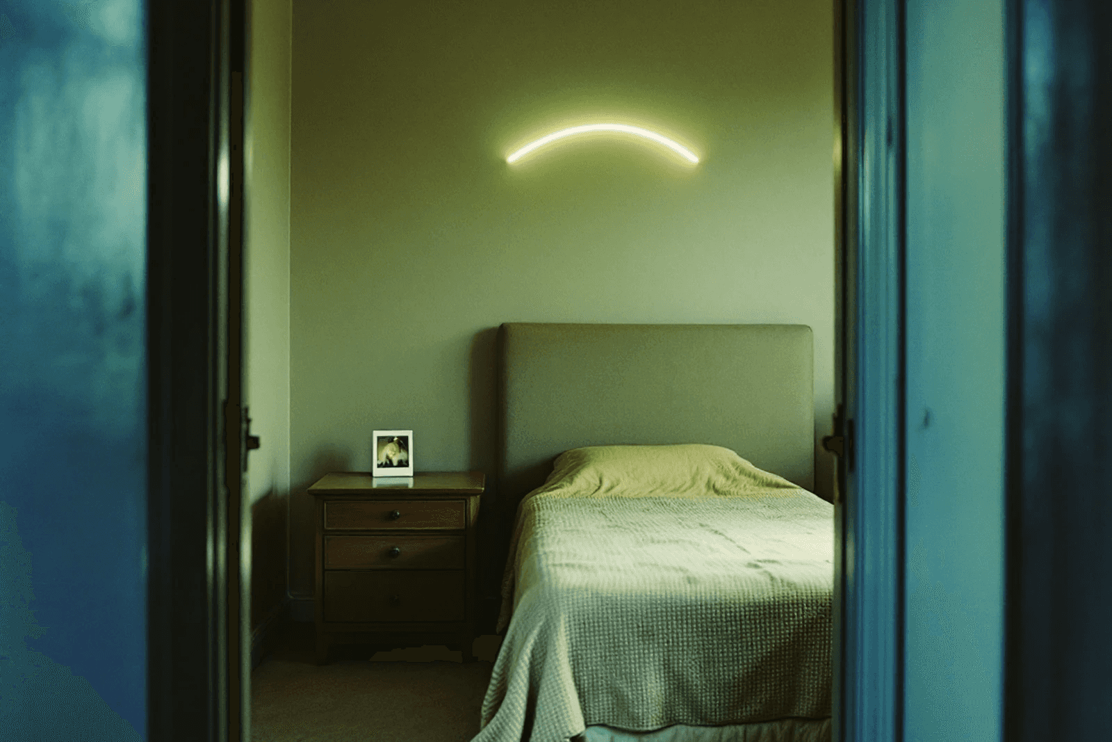 A Polaroid print face down on a bedside table in an empty 1970s bedroom, a pale arc of light on the wall.