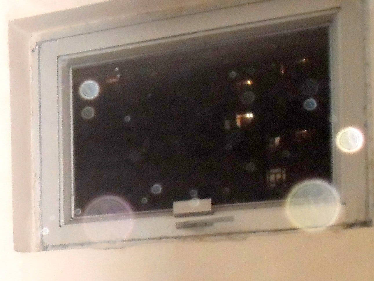 A small window in a pale wall at night, with dark buildings and a few lit windows outside, and translucent circles of different sizes over the glass and the frame.